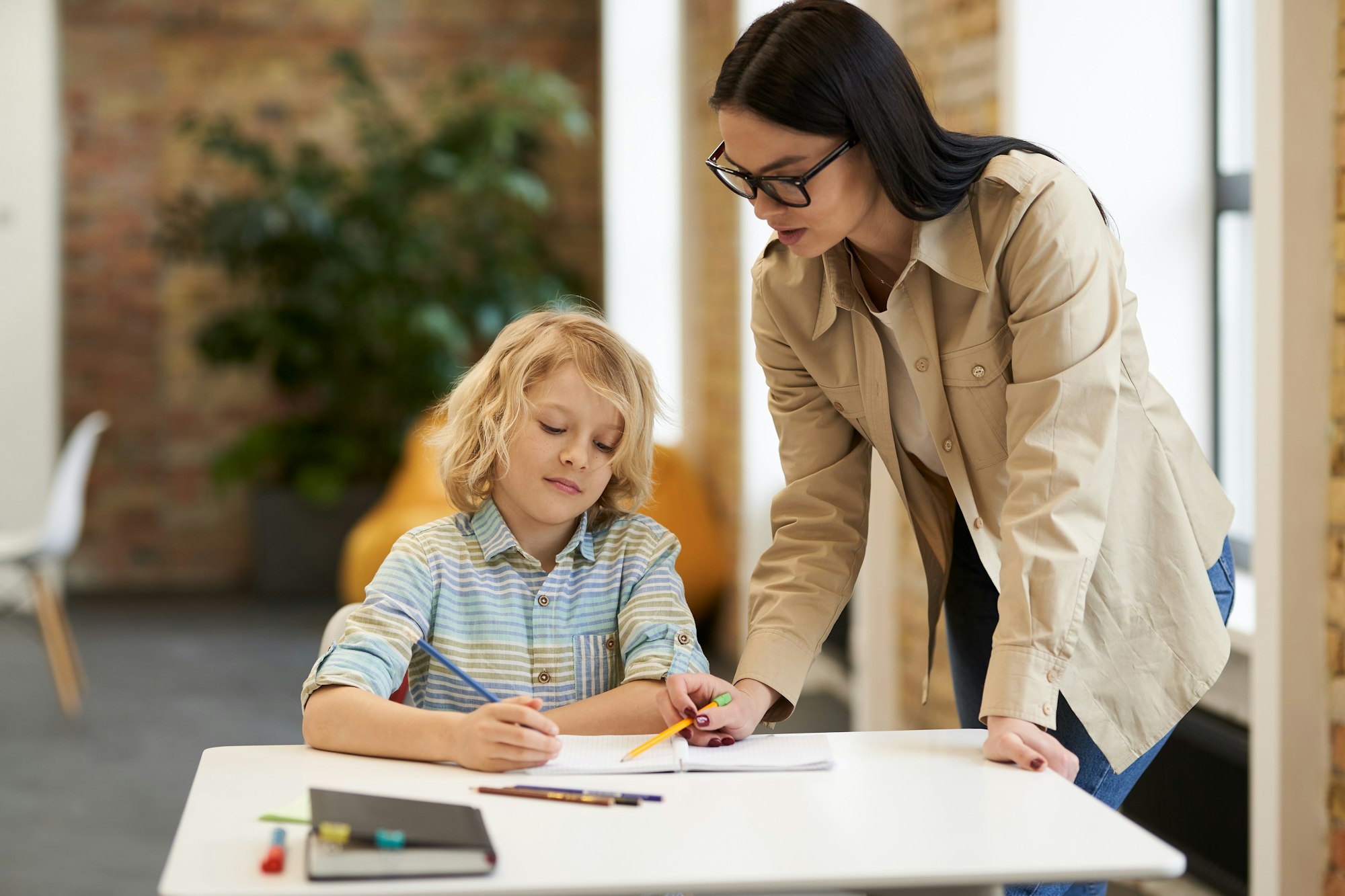 Supportive young female teacher in glasses helping little boy with the task. Kid studying in